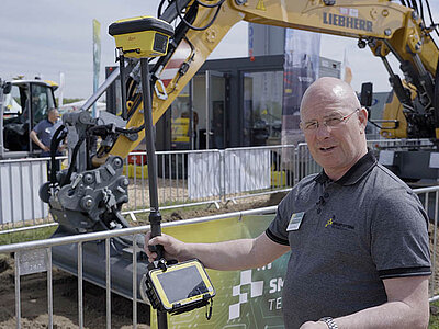 Mitarbeiter von Smart Systems erklärt einen Rover-Stab im Hintergrund ein Liebherr Bagger