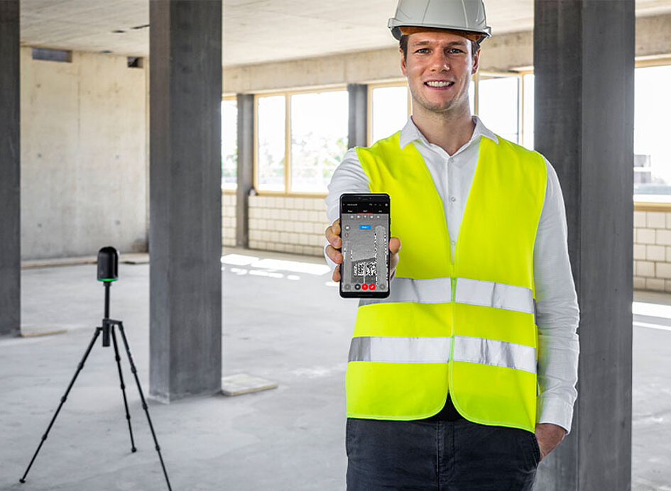 Arbeiter in Warnweste hält ein BLK 3D in die Kamera. Im Hintergrund zu sehen ein BLK2Go Gerät