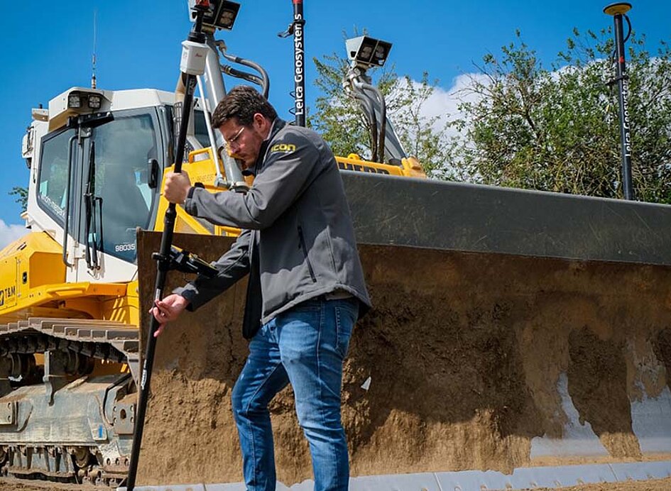 Vermessung einer Baustelle mit einem AP20 Autopole von Leica. Im Hintergrund eine Raupe mit Maschinensteuerung.