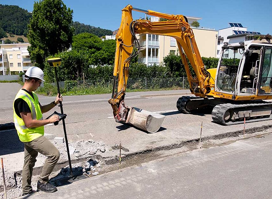 Straßenbau mit Raupenbagger und Arbeiter der mit GNSS-Rover iCG160 die Baustelle vermisst