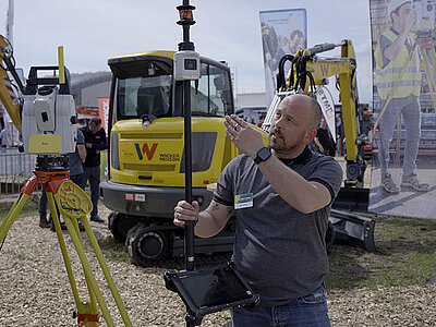 Ein Smart Systems Mitarbeiter erklärt einen AutoPole Vermessungsstab im Hintergrund weitere Vermessungsgeräte und Bagger