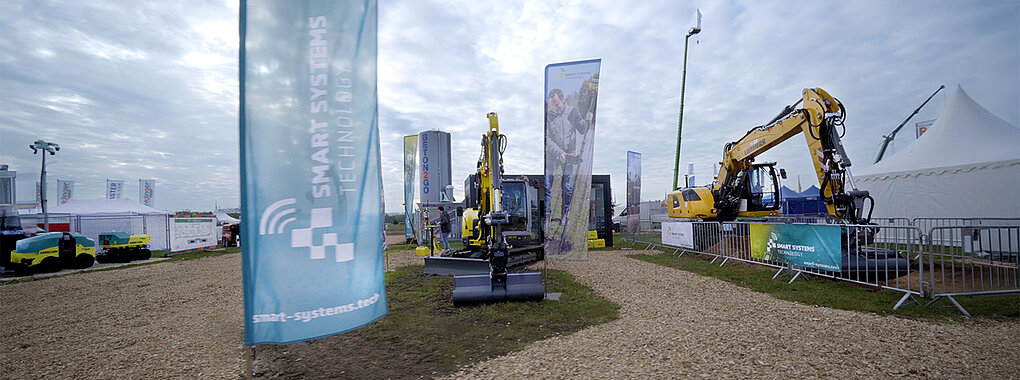 Bild zeigt Bagger und Flaggen von Smart Systems Technology auf der Messe TiefbauLive im Jahr 2023