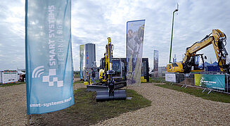 Bild zeigt Bagger und Flaggen von Smart Systems Technology auf der Messe TiefbauLive im Jahr 2023